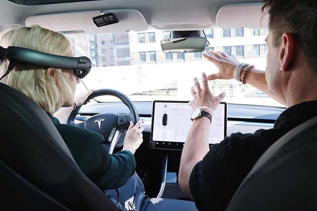 A driver in a Tesla wearing digital headgear, while the passenger gestures to something in front of the car.