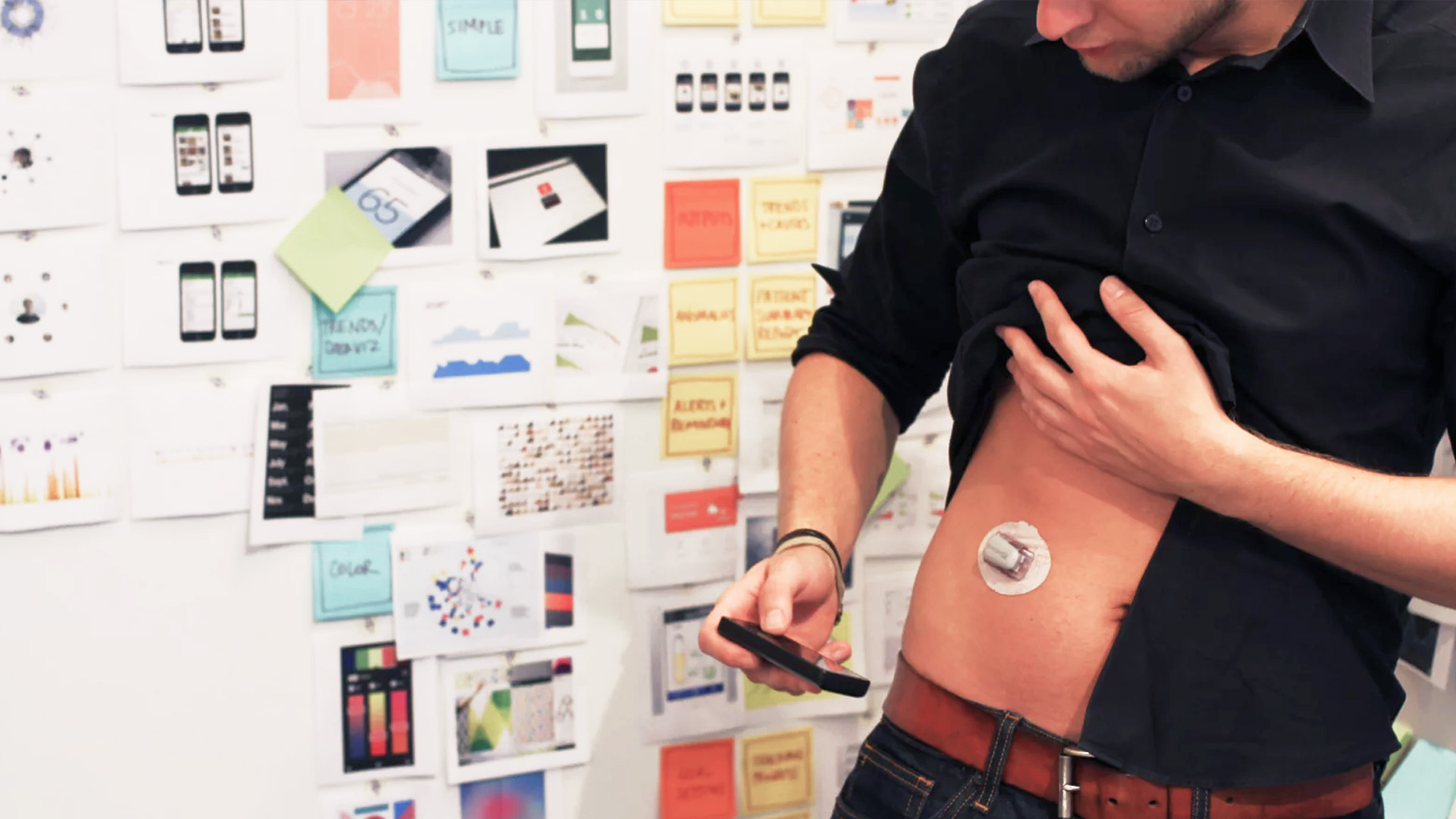 An IA Collaborative designer with stick-on medical probe attached to his abdomen, looking at smartphone app.