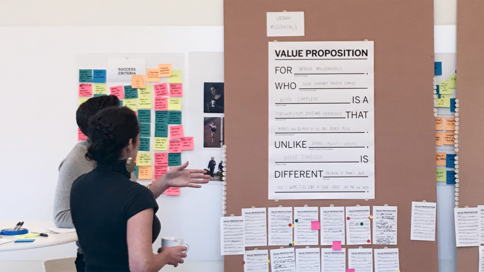 Two IA Collaborative designers standing in front of a wall of written notes, looking at large poster entitled "Value Proposition".