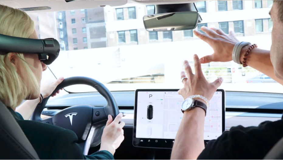 A driver in a Tesla wearing electronic headgear while the passenger gestures toward the front of the car.