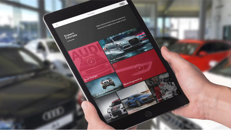Two hands holding a tablet displaying the MyAudi app, with an Audi dealership in the background.